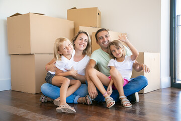 Fototapeta na wymiar Smiling family with kids sitting on floor near cardboard boxes and relaxing. Blonde girl on father legs waving. New home owners enjoying apartment. Mortgage, relocation and moving day concept