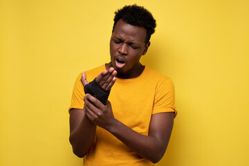 Young african american man holding his hand having pain in wrist. Studio shot on yellow wall.