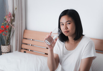 Beautiful Asian woman drinking fresh water from glass in white bedroom interior background