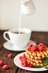 closeup view of delicious belgian waffles with raspberries and mint leaves on wooden table, cup of coffee on background 