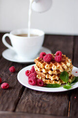 closeup view of delicious belgian waffles with raspberries and mint leaves on wooden table, cup of coffee on background 