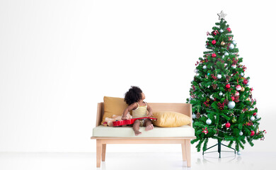 Obraz na płótnie Canvas Dark skinned little girl sitting on bench and looking at Christmas tree, white background