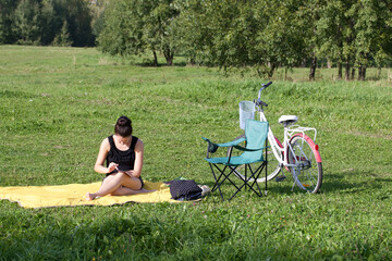 The girl is resting in the park. Sits on the rug, looks at the phone. There is a bicycle and a camping chair nearby.