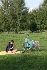Obraz premium The girl is resting in the park. Sits on the rug, looks at the phone. There is a bicycle and a camping chair nearby.