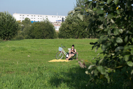 The girl is resting in the park. Sits in a camping chair, looks at the phone. There is a bicycle nearby.