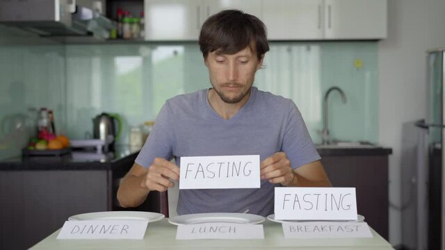 A happy young man sits in front of a table. He puts signs fasting on an empty plates with signs breakfast, lunch, and dinner. Interval fasting concept. Skipping meals
