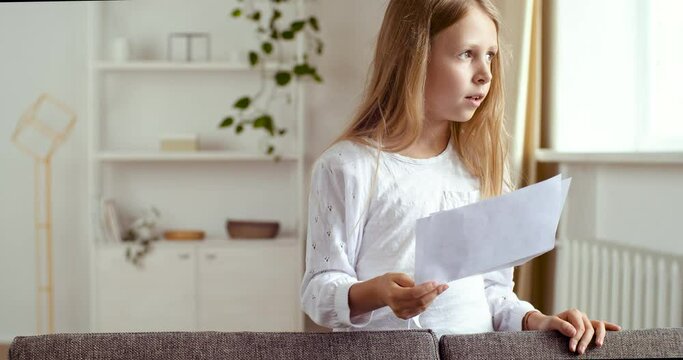 Focused girl of 7-10 years old stands at home reading script of play, preparing for casting for role in school theater. Lady small child repeats rules, learns verse, does homework in room in qurantine