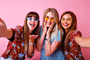 Studio portrait of three super exited best friends girls, blissful positive vibes, summer bright tropical print trendy clothes and accessories, pink background, sisters having fun.