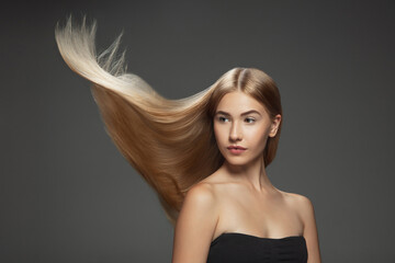 Beautiful model with long smooth, flying blonde hair isolated on dark grey studio background. Young caucasian model with well-kept skin and hair blowing on air. Concept of salon care, beauty, fashion.