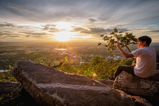 man sitting relax and holding a cellphone taking pictures on top of a moutain watching the sunset