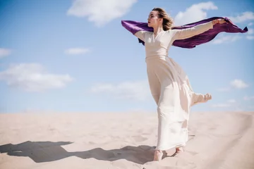 A young, slender girl in a beige dress with purple cloth in her hands posing in the desert in the wind © Denis