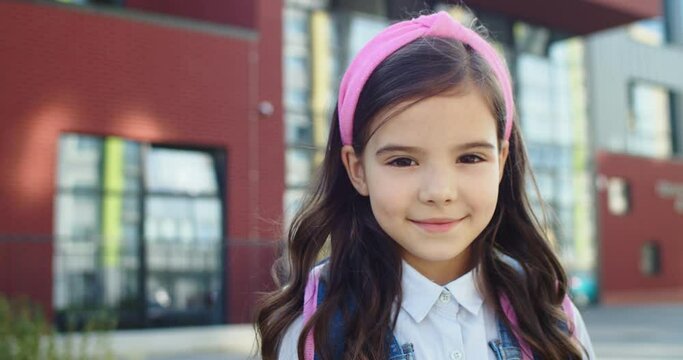 Close up portrait of adorable beautiful Caucasian little girl smiling in front of school. Pretty cute school kid standing with smile on face outdoors alone. Back to school. Pupils concept