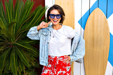 Summer spring bright positive portrait of happy smiling brunette woman wearing trendy feminine hipster outfit smiling and having fun, posing in front of surf boards and palms.