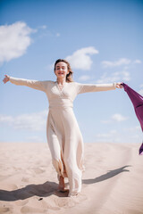 A young, slender girl in a beige dress with purple cloth in her hands posing in the desert in the wind