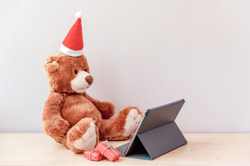 Funny teddy bear Santa in Christmas hat with gifts sitting at the tablet. Photography for ad of toy online store