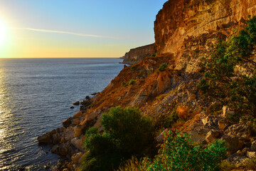 High red brown yellow sandy long cliffs of Fiolent with green bushes on the Black Sea coast in the light of sunset. Blue water and sky. Gradient on horizon. South Crimea