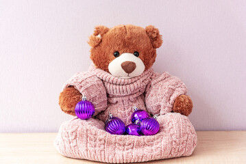 Cute teddy bear in knitted sweater with little lilac Christmas balls. New year greeting postcard