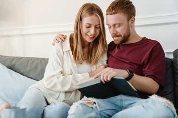 Photo of happy couple hugging and reading book while sitting on couch