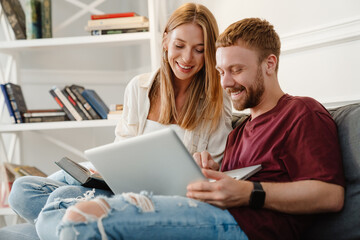 Image of couple reading book and using laptop while sitting on sofa