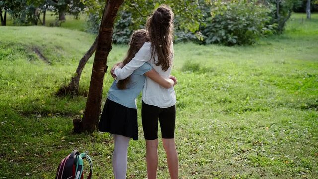 Children hug in the park and look ahead. Back view of two sisters of different ages and heights, in different styles of clothing