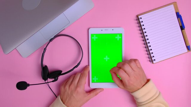 Top view - Workplace of person from online technical support. Hands of consultant using digital table with green screen on pink background. Male hands scrolling pages, tapping on touch screen.