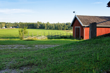 Obraz premium Traditional Finnish red wooden barn with roundpole fence and the weather vane on the roof. Old idyllic farm in the countryside.