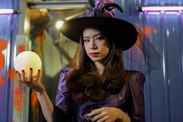 Asian young girl dressed as a witch holding a magic crystal ball in hand and casting a spell on...