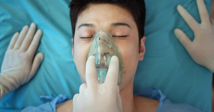 Close up of unconscious asian man with oxygen inhalation mask dying while lying on medical stretcher. Paramedics checking pulse to millennial guy while they going to operation room