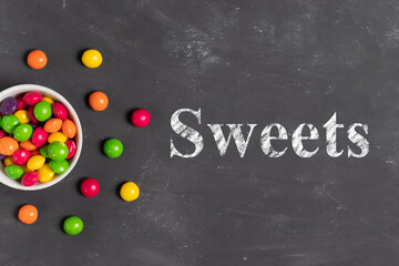 Multicolored round candies in a white plate on a gray board with the inscription sweets