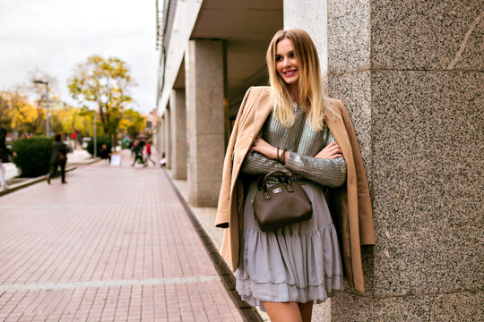 Fashion street style picture of blonde elegant woman, wearing luxury silk dress, trendy sweater, cashmere coat and leather bag, soft warm colors, spring autumn mid season mood.