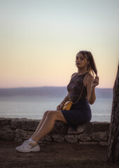 Fototapeta na wymiar Attractive brunette sitting posing on a small stone wall, late sunset in the afternoon. Moving her hands sensually, dressed in blue skirt with a yellow purse.