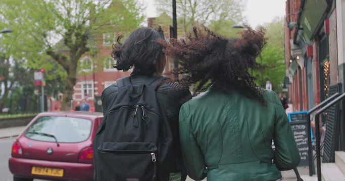 Two woman walk down the street together as wind blows their hair.