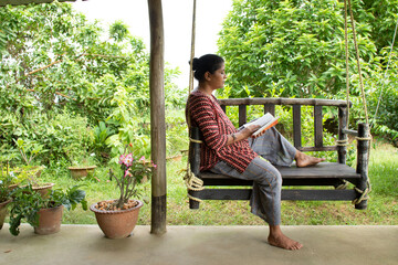 Bolpur, West Bengal/India - 09.13.2020: A pretty woman sitting on a wooden swing reading a book in a beautiful environment