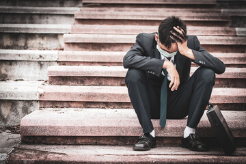 Failure unemployed stressed young asian business man in suit and wearing a face mask covering head with hands. A young businessman sitting desperate on the stairs because low economic crisis.