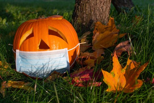 Carved Halloween pumpkin with sad eyes wearing mask on the grass with leaves, no party during Covid or Coronavirus outbreak, events and parties canceled to prevent the risk of spreading the virus