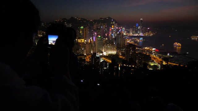 Man take long exposure pictures of night Hong Kong from top of hill. Dark blurred silhouette on foreground, colorful city panorama on background. Beautiful views of Asian metropolis
