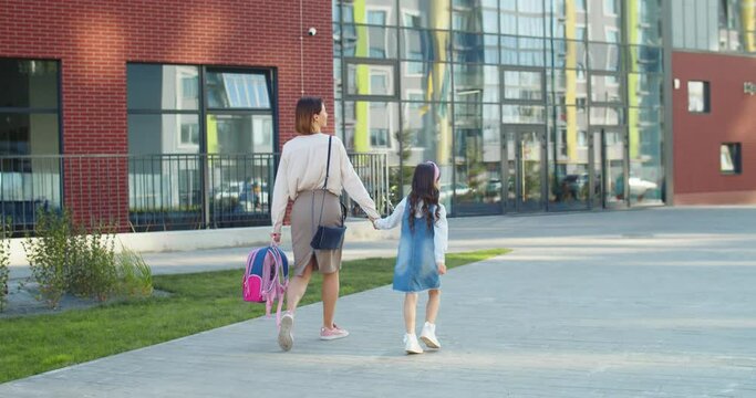 Rear of beautiful Caucasian woman walking with little daughter junior student to school and speaking on sunny day. Mother leads girl to school and talking while holding pink backpack Education concept