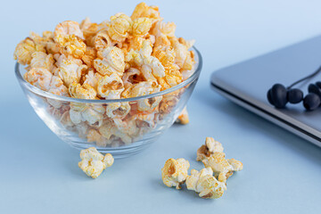 A transparent bowl of popcorn on a blue surface. Next to a laptop and headphones. Home family watching movies.