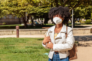 Young African American woman with curly hair and face mask walks holding several books outdoors on a very sunny day. College or high school student.