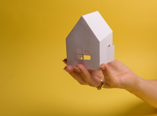 White family paper house in man hand on yellow background paper. Minimalistic style. Copy space. View from above. Vertical orientation.