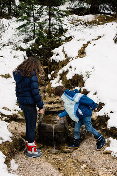 Kids playing, boy puts a snowball into the crystal clear mountain stream in Winter