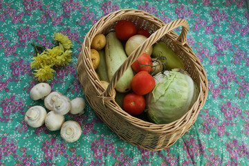 A basket with an autumn harvest of vegetables against the background of a flowered tablecloth. The basket contains fresh vegetables and mushrooms