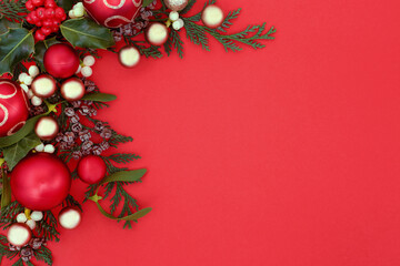 Christmas background border with bauble decorations, holly & winter greenery of mistletoe, ivy, & cedar cypress fir on red. Xmas New Year composition for the festive season. Top view, copy space.