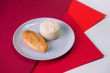 Side view of Tasty roasted chicken fillet on gray plate with boiled white rice served in culinary ring. Breakfast on colorful red background. Main and side dish in one plate.
