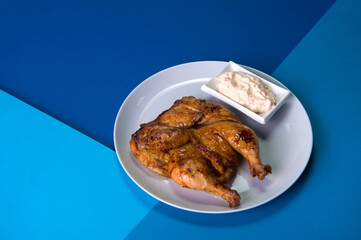 Grilled chicken Tabaka on gray plate served with sauce in white bowl. Georgian cuisine. Restaurant menu food on blue abstract background 
