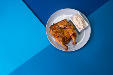 Grilled chicken Tabaka on gray plate served with sauce in white bowl. Georgian cuisine. Restaurant menu food on blue abstract background 

