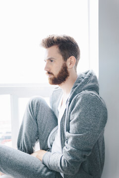 Man in a grey hoodie and sweat pants sits at a window