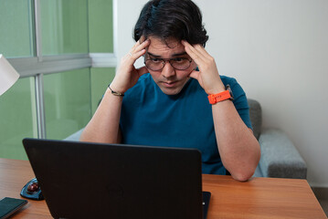 Man worried, tired or stressed looking at his laptop, holding his head with his hands, in a wooden desk at home