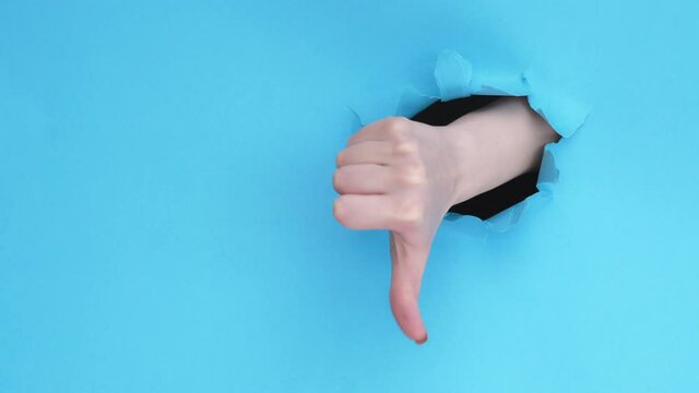Vote gesture. Breakthrough paper hole. Female hand showing yes no thumb up down isolated on blue ripped wall background with copy space. Changing mind. Rejection sign. Approval dislike loop.