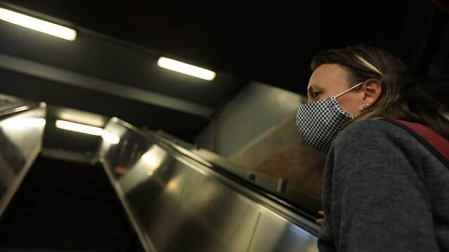 Young woman wearing coronavirus face protection mask standing with hand on rails going upstairs on escalator waiting to reach upwards